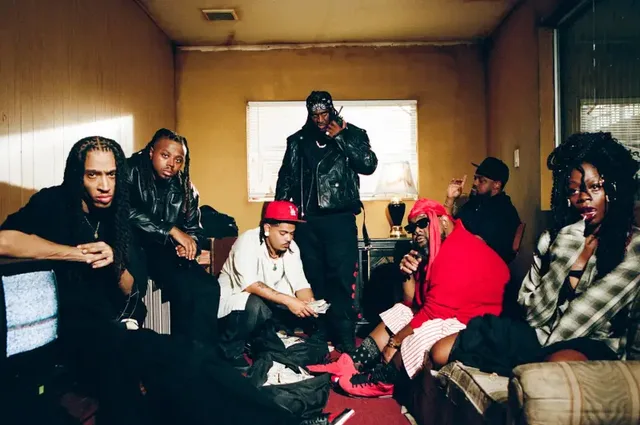 Eight people together in a wood-panelled room, some seated on the floor and a sofa, one standing in a leather jacket and bandana.