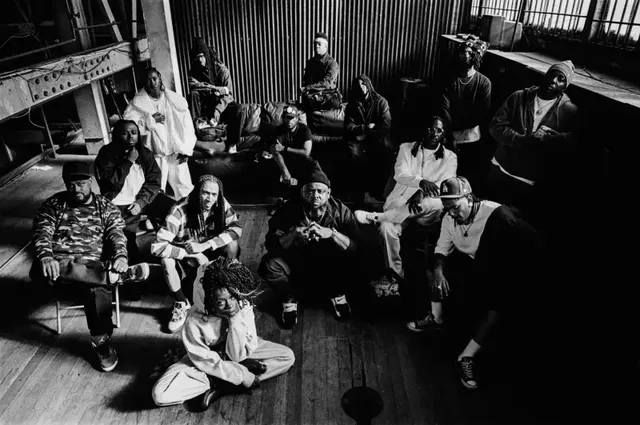 A large group photographed from above in an industrial space: corrugated metal walls and a steel beam overhead, people seated along a ledge, on crates and on the wooden floor, one person sitting cross-legged at the front. Black and white.